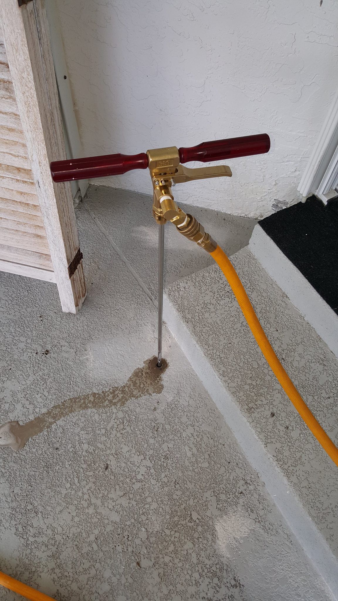 A brass water-wand sprayer with a red grip attached to a yellow garden hose on a concrete patio.