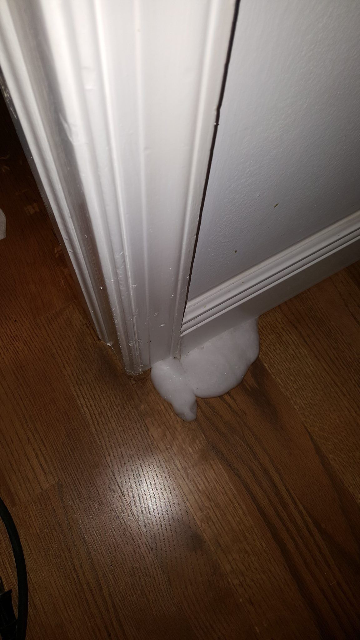 A pile of white spray foam insulation leaking from the base of a white door frame onto a wooden floor.