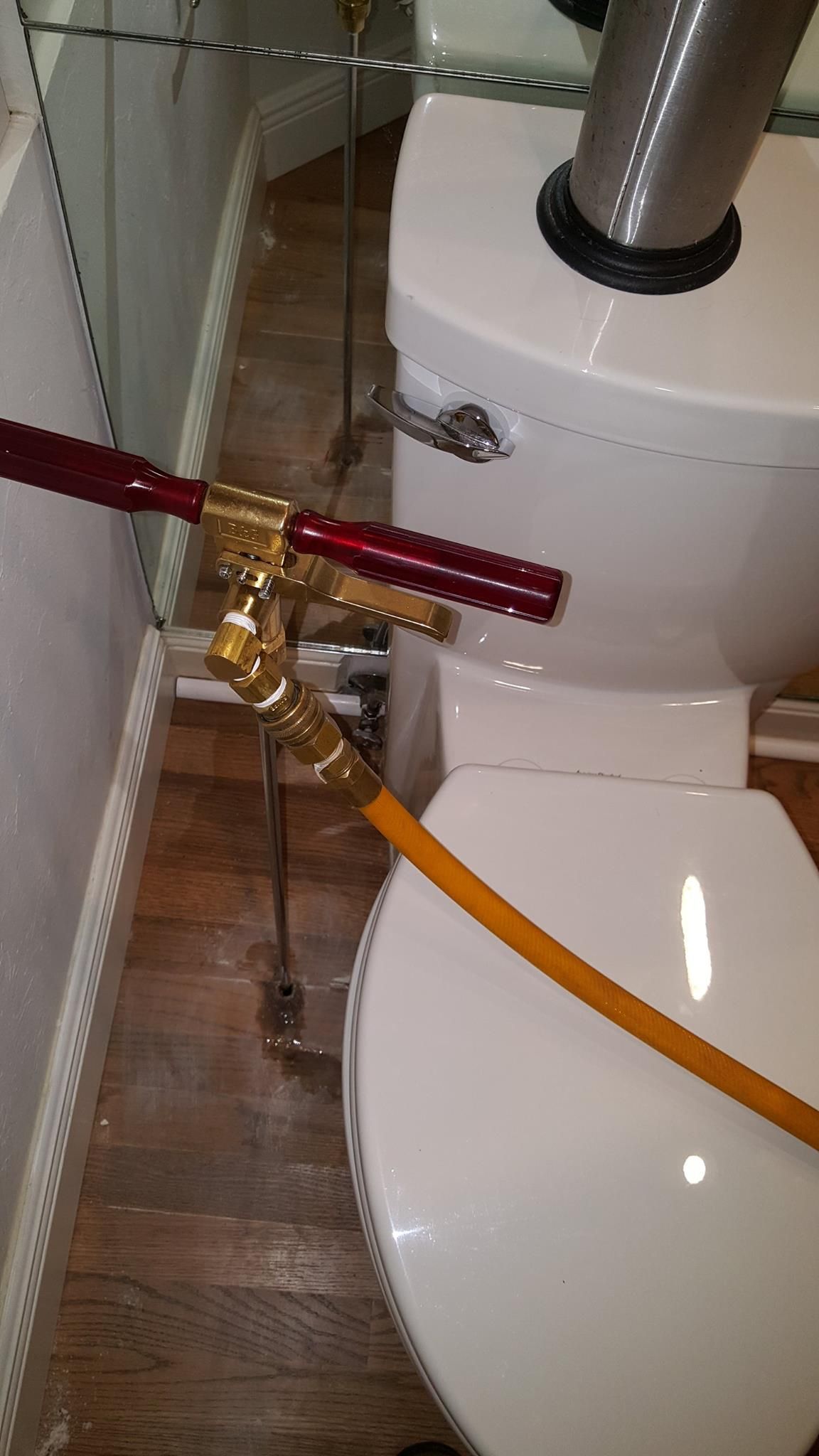 A brass-handled, orange-tubed reach-in toilet tool positioned next to a white toilet in a bathroom.