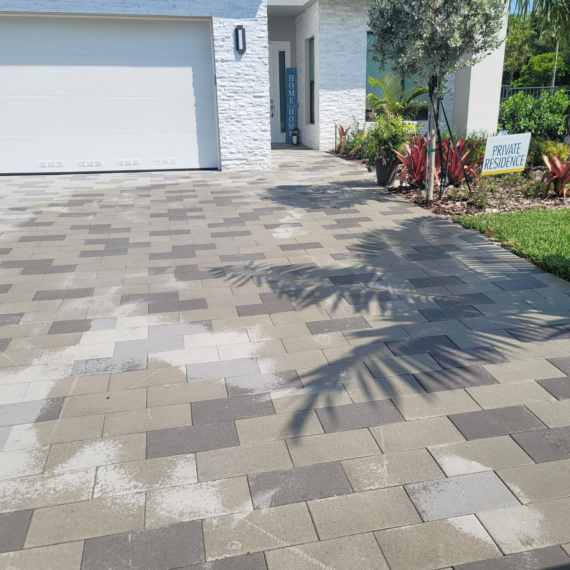 A paved driveway made of gray and tan rectangular pavers leading to a modern house with a white stone facade.