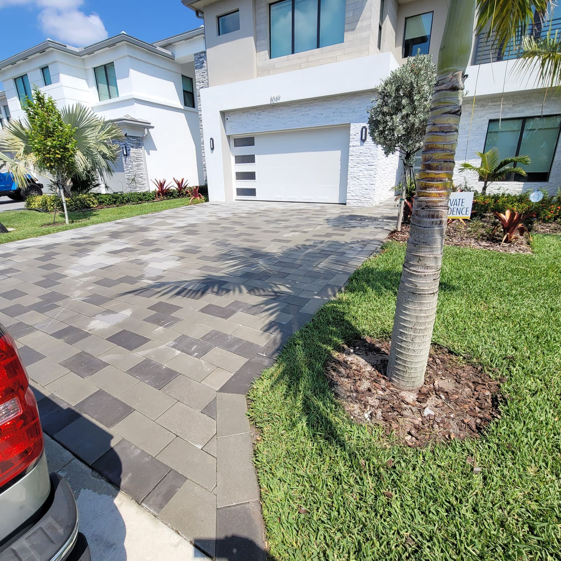 A modern two-story home with a paved driveway, a palm tree in the foreground, and lush green landscaping.