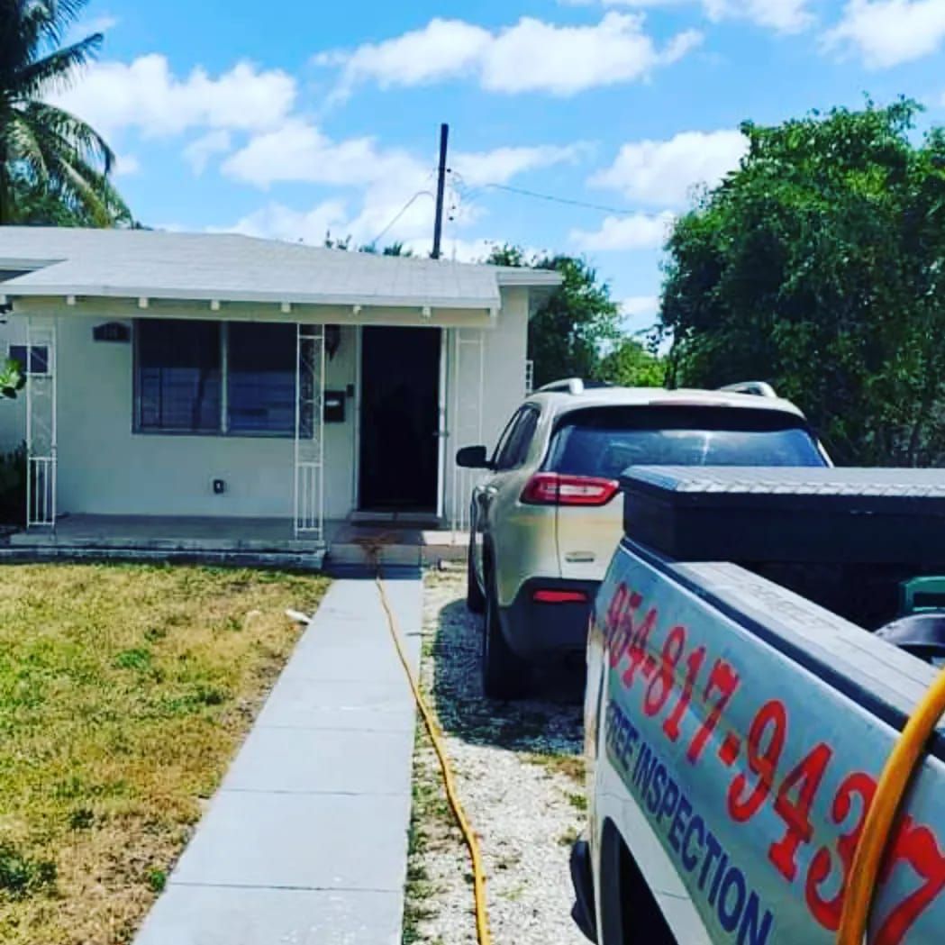 A single-story white house with a walkway, a car parked in the driveway, and the back of a utility truck with contact info.
