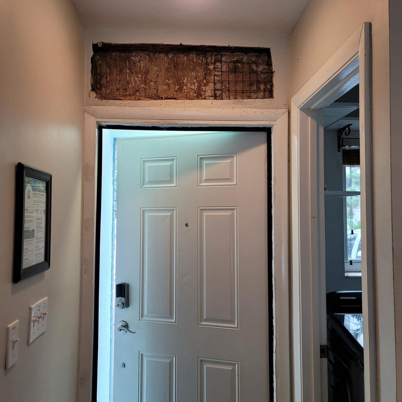 Interior view of a white door with a patch of wall above it removed, exposing the underlying lath and plaster structure.