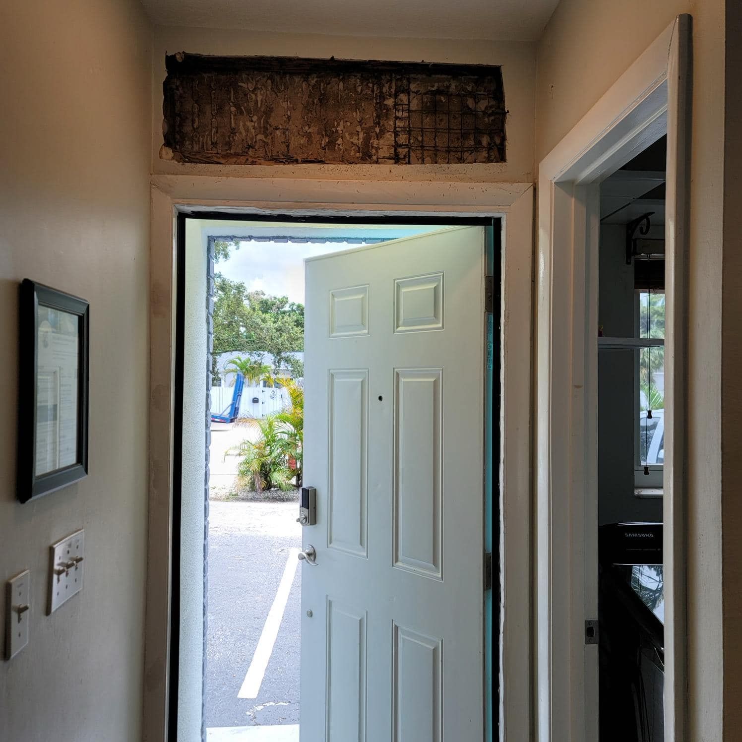 An open white door reveals an outdoor parking lot. Above the door frame is an unfinished rectangular wall opening.