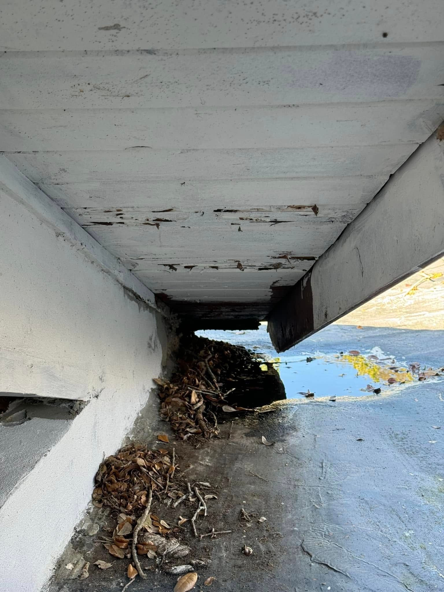 A close-up view of a concrete bridge underside showing peeling paint, rusted structural joints, and debris on the ground.