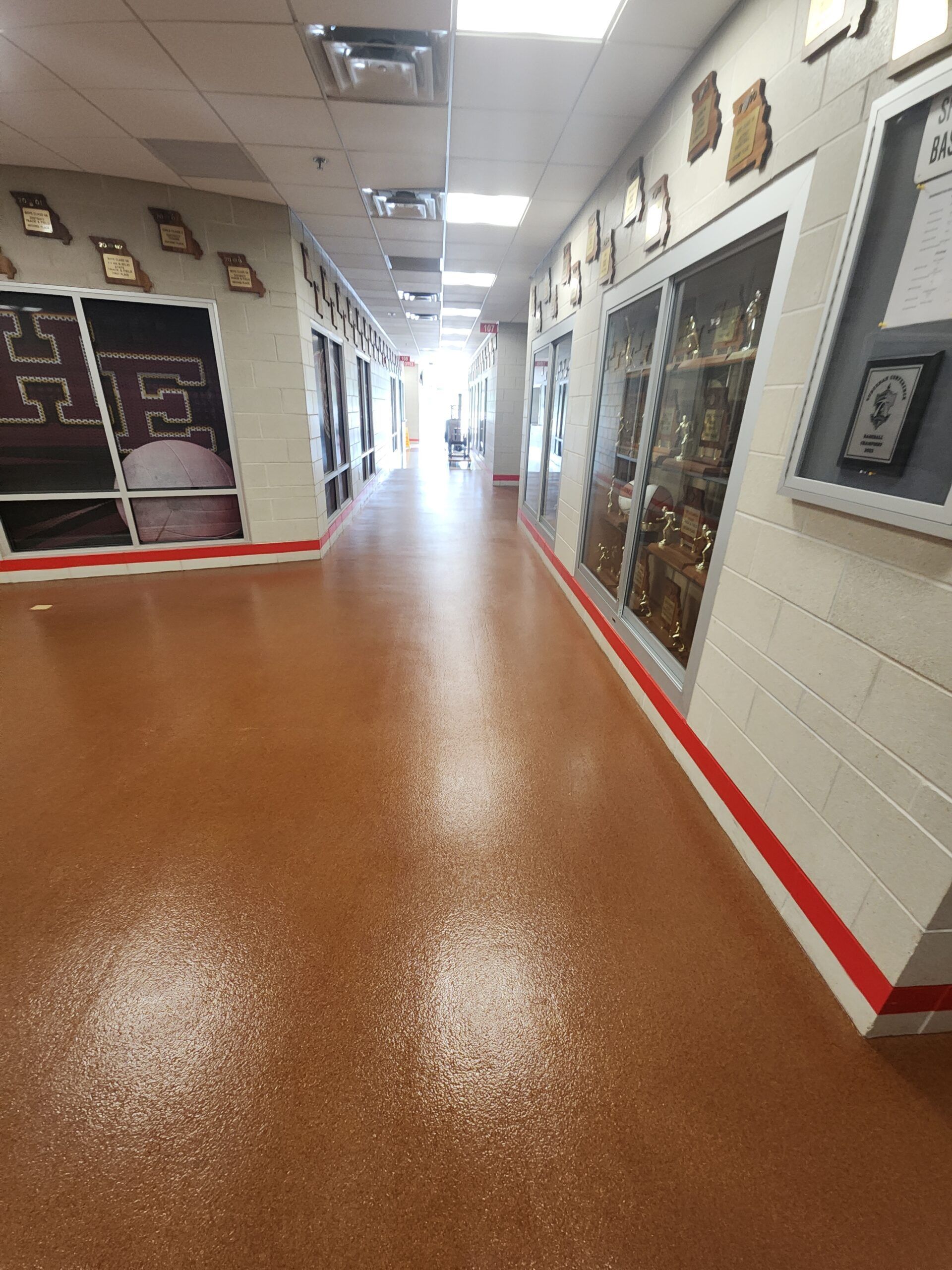 A hallway with a speckled brown floor, red baseboards, and walls lined with trophy cases and framed awards.