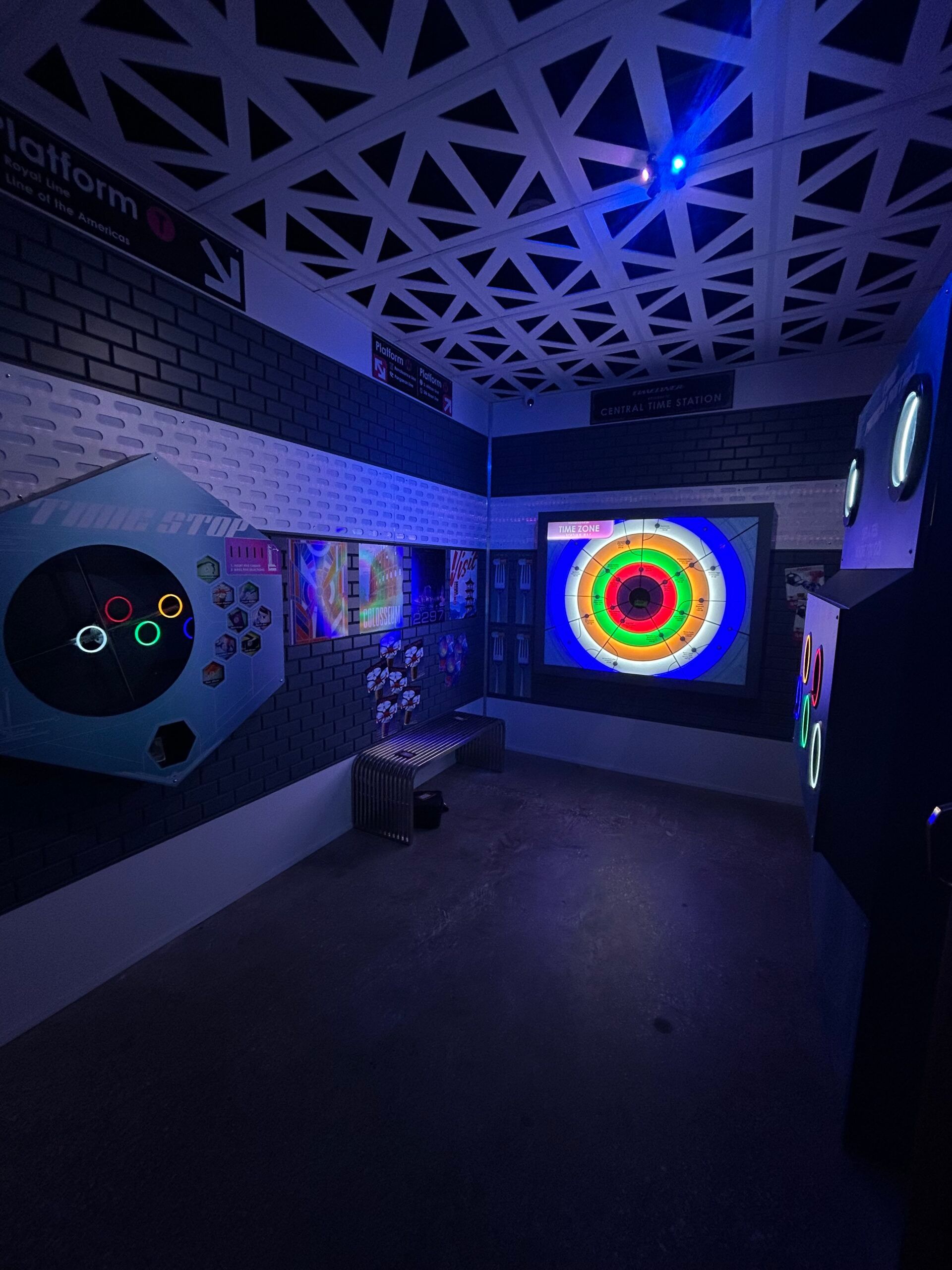 A dimly lit gaming area features a colorful digital target on a wall, a geometric ceiling pattern, and station signage.