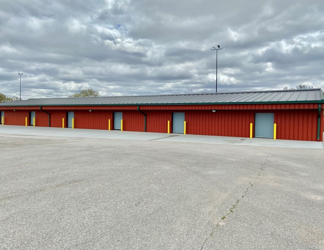 A long, single-story red storage building with multiple grey metal doors, situated in a large, paved parking lot.