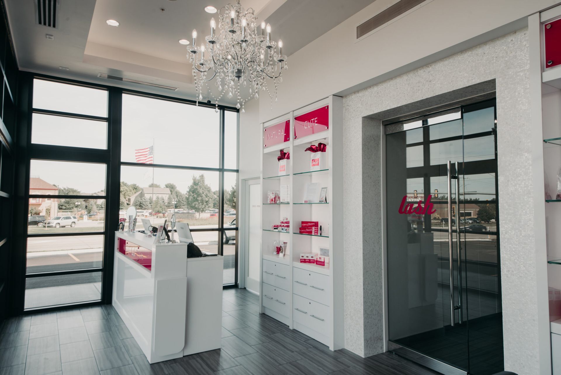 A white reception area with a glass chandelier, large windows, sleek displays, and a glass-paneled office door.