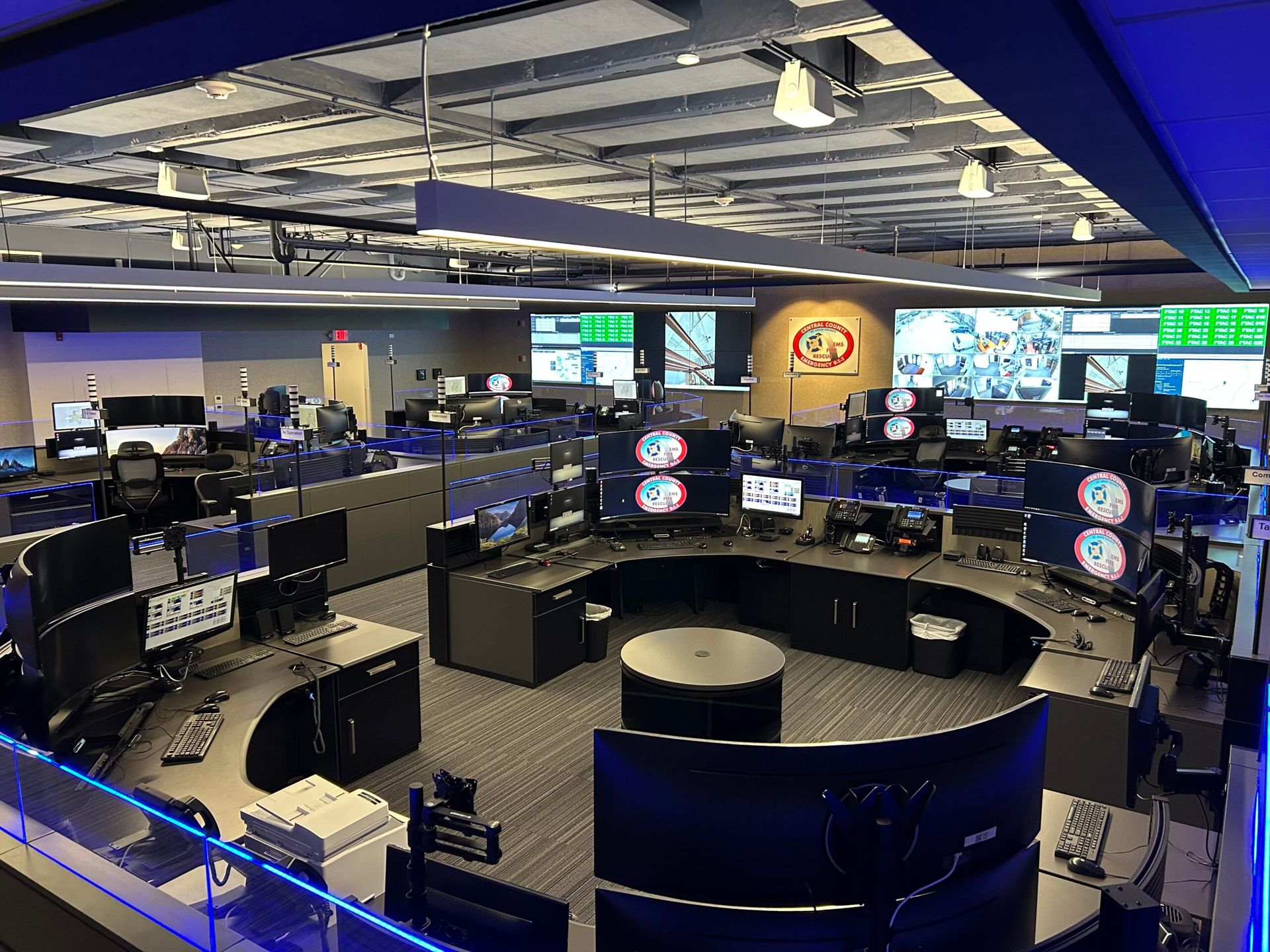 A modern, curved control room with multiple computer workstations, wall-mounted monitors, and blue accent lighting.