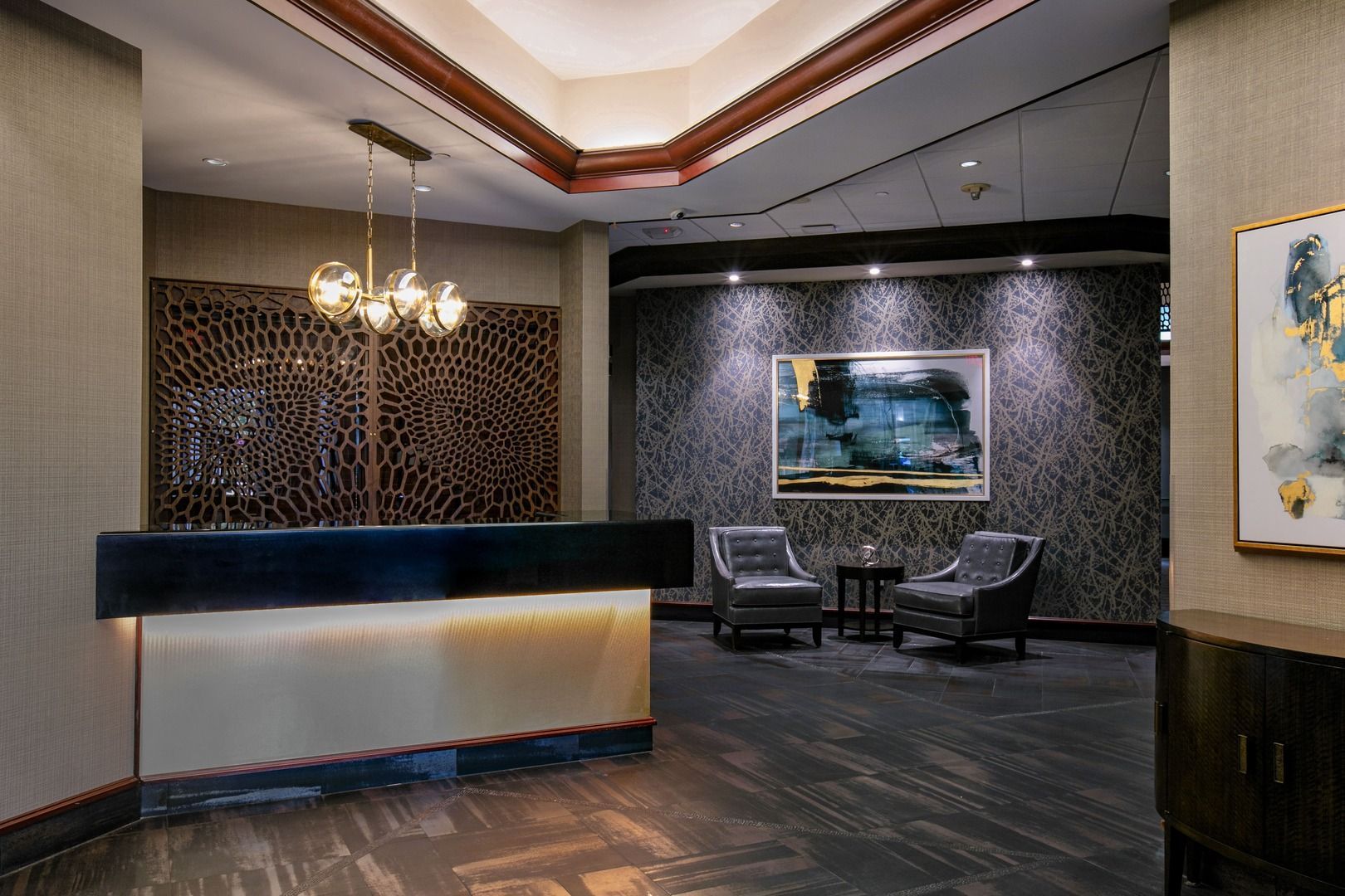 A hotel reception area with a lit desk, modern pendant lighting, patterned wall panels, two armchairs, and wall art.