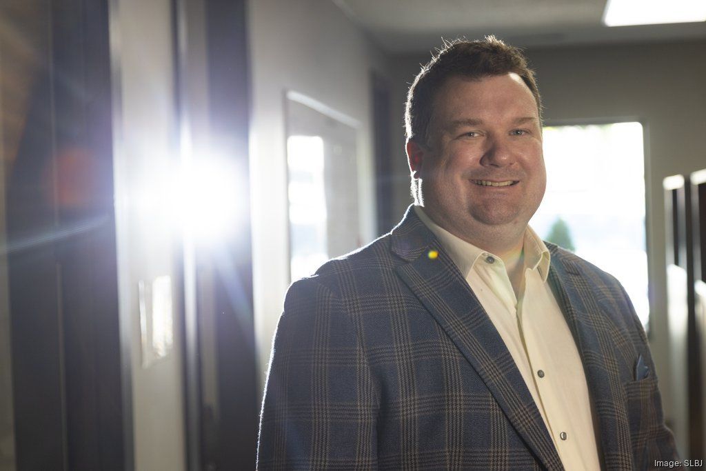 A person wearing a plaid blazer and light shirt smiles in a sunlit hallway with strong lens flare.