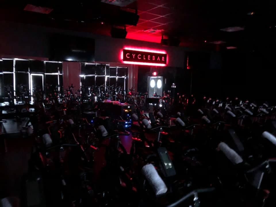 A dark CycleBar fitness studio interior with rows of stationary exercise bikes facing a glowing red neon sign.