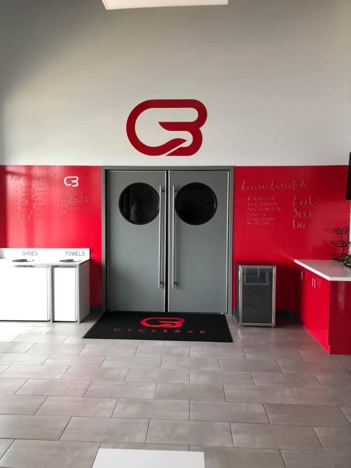 A gray double door centered beneath a red brand logo on a red wall, with a black branded floor mat and tiled flooring.