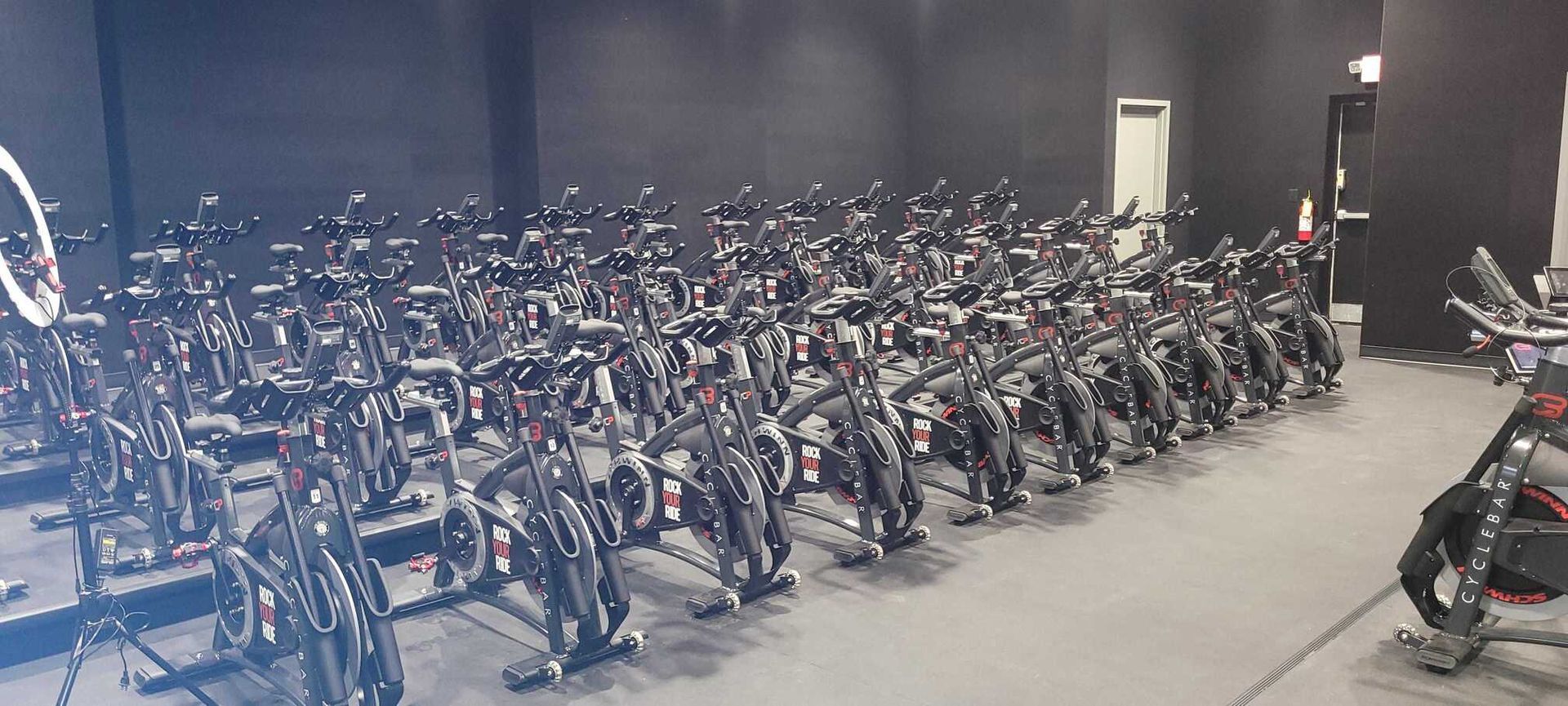Rows of black stationary cycling bikes lined up in a gym with dark walls and a gray floor.