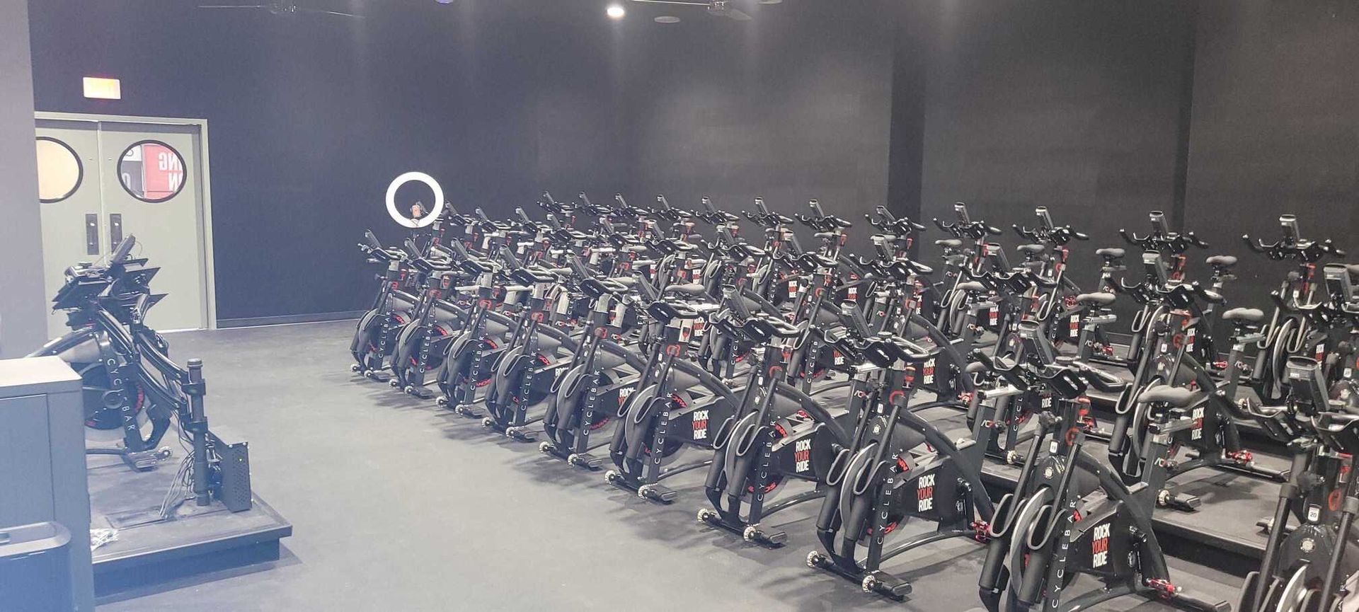 A studio filled with rows of black stationary bikes arranged for a group cycling class against a dark, minimalist wall.