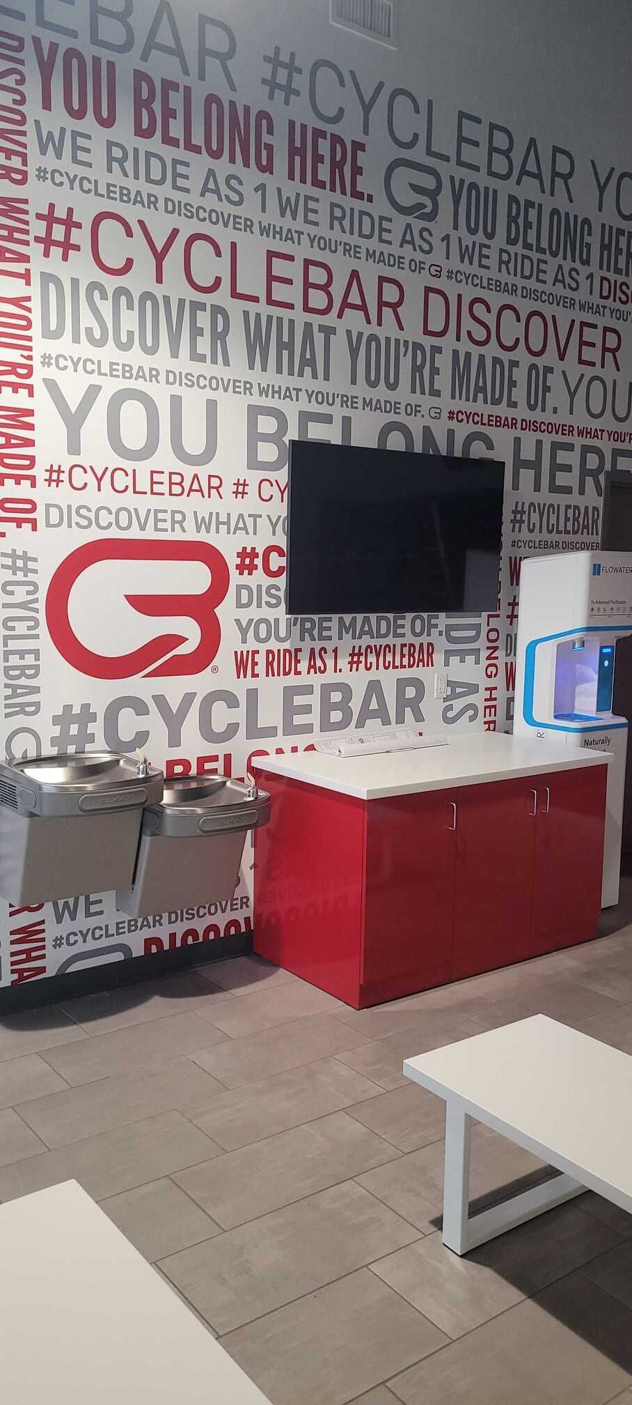 A branded CycleBar wall featuring motivational text and logos behind a red desk, a water fountain, and white tables.