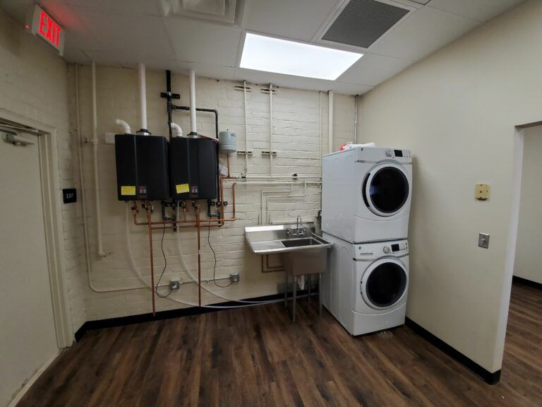 A laundry room with two stacked white washers/dryers, a stainless steel sink, and two wall-mounted black water heaters.