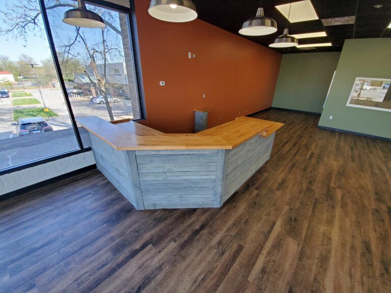 A wooden reception desk with a gray plank front sits in an office with orange walls, wood-look flooring, and large windows.