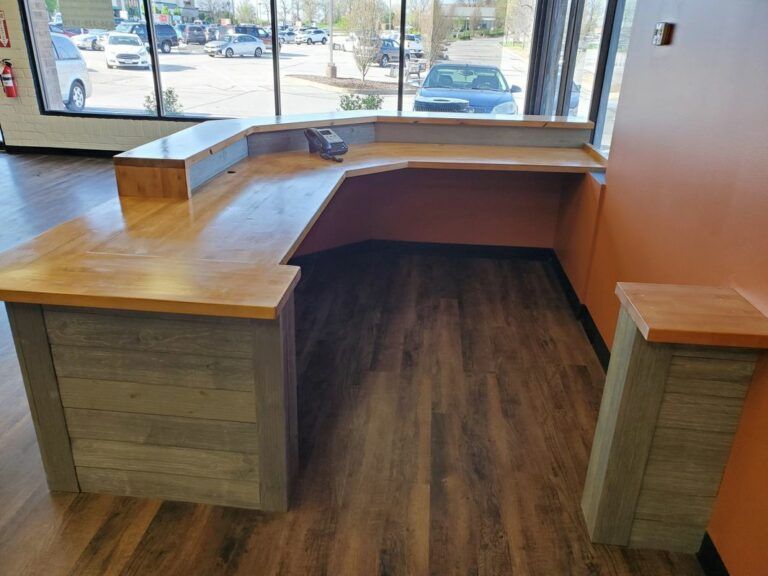 An L-shaped reception desk with a wood top and gray plank base, situated in an office by large windows.