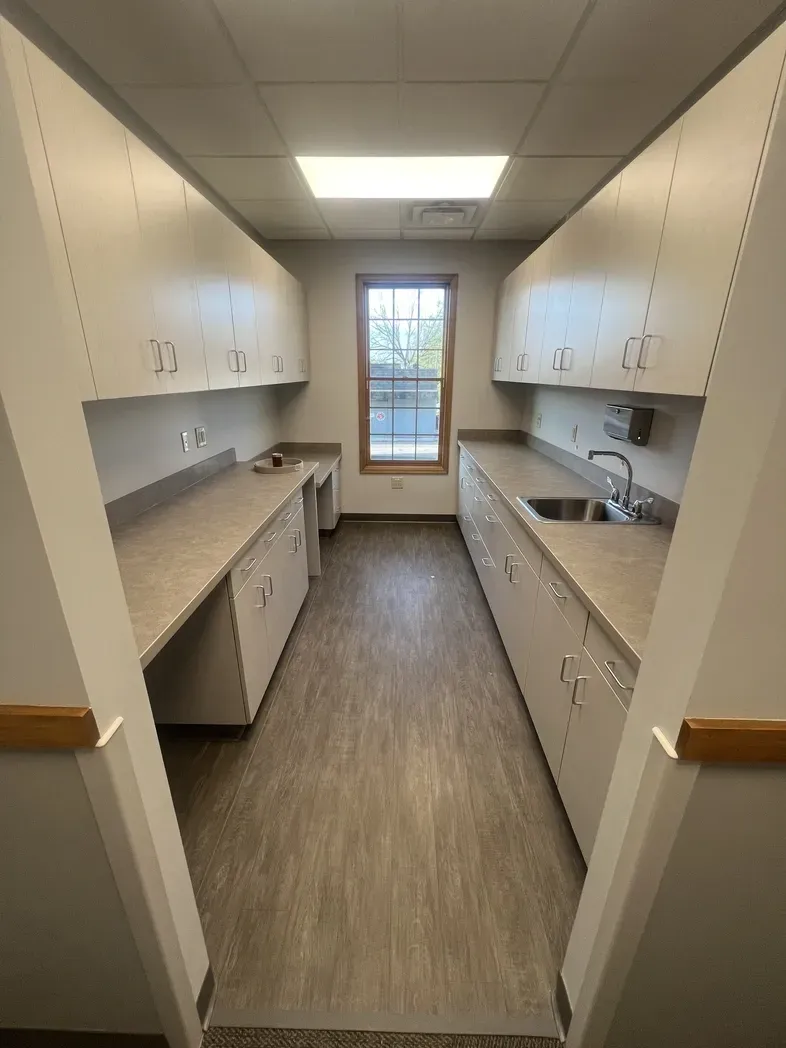 A galley-style utility room with beige cabinets, light-colored countertops, a sink, and a window at the end.