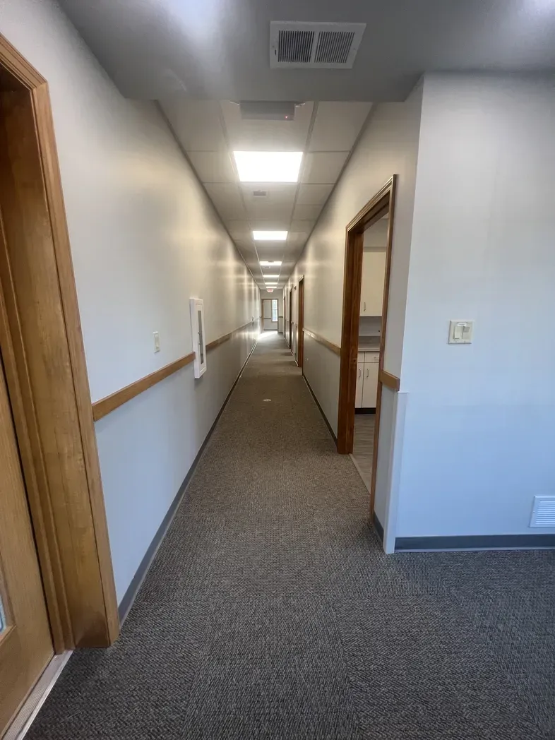 A long, straight office hallway with neutral walls, light brown trim, grey carpet, and recessed ceiling lights.