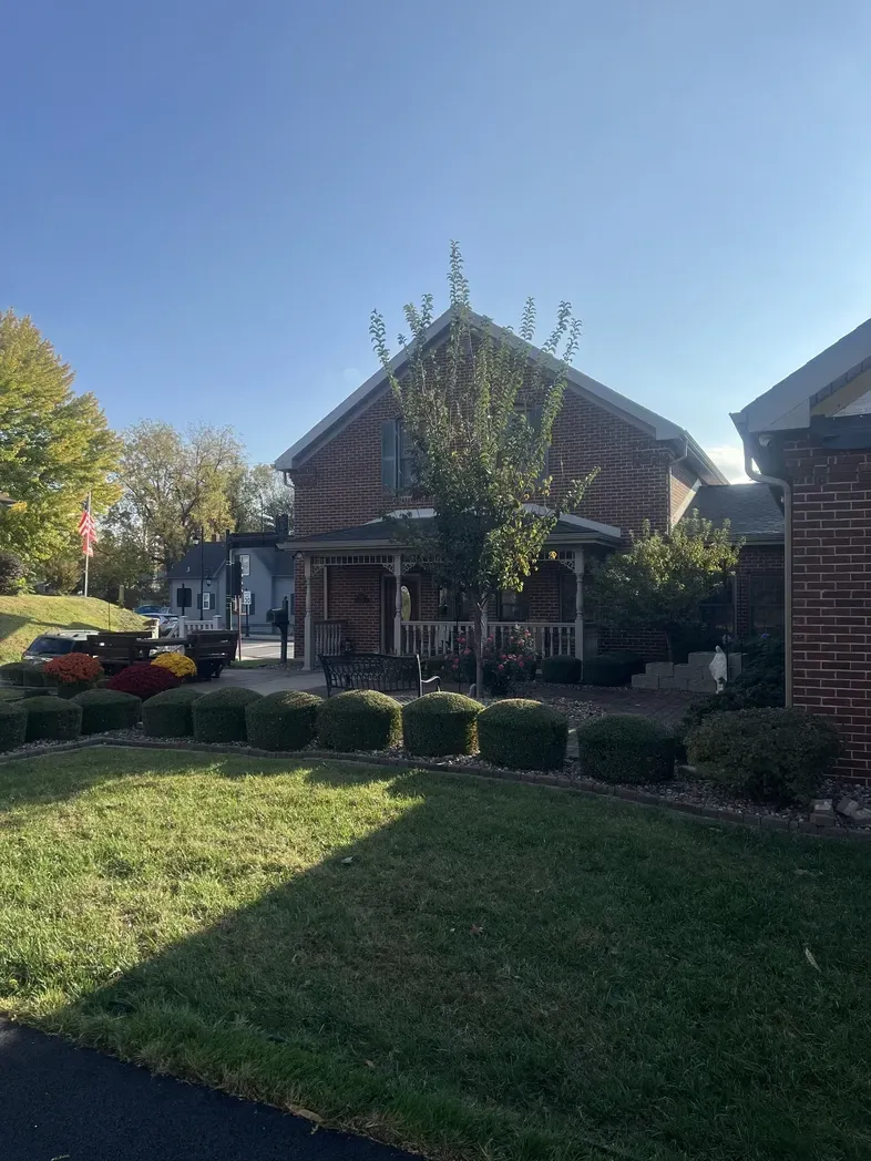 A brick building with a front porch, surrounded by manicured shrubs, a green lawn, and trees under a clear blue sky.
