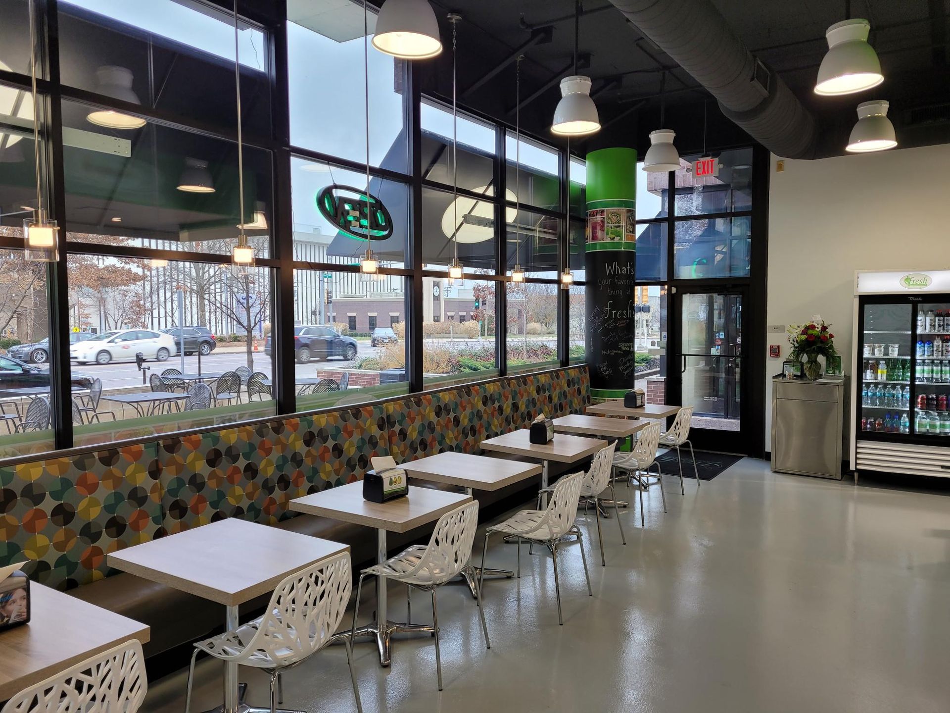 An empty dining area with small square tables, white patterned chairs, a long bench, and overhead pendant lighting.