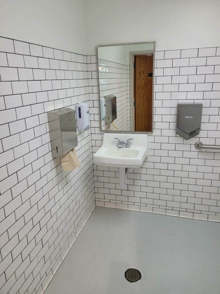 A white-tiled bathroom corner with a sink, mirror, wall-mounted paper towel dispenser, and hand dryer.