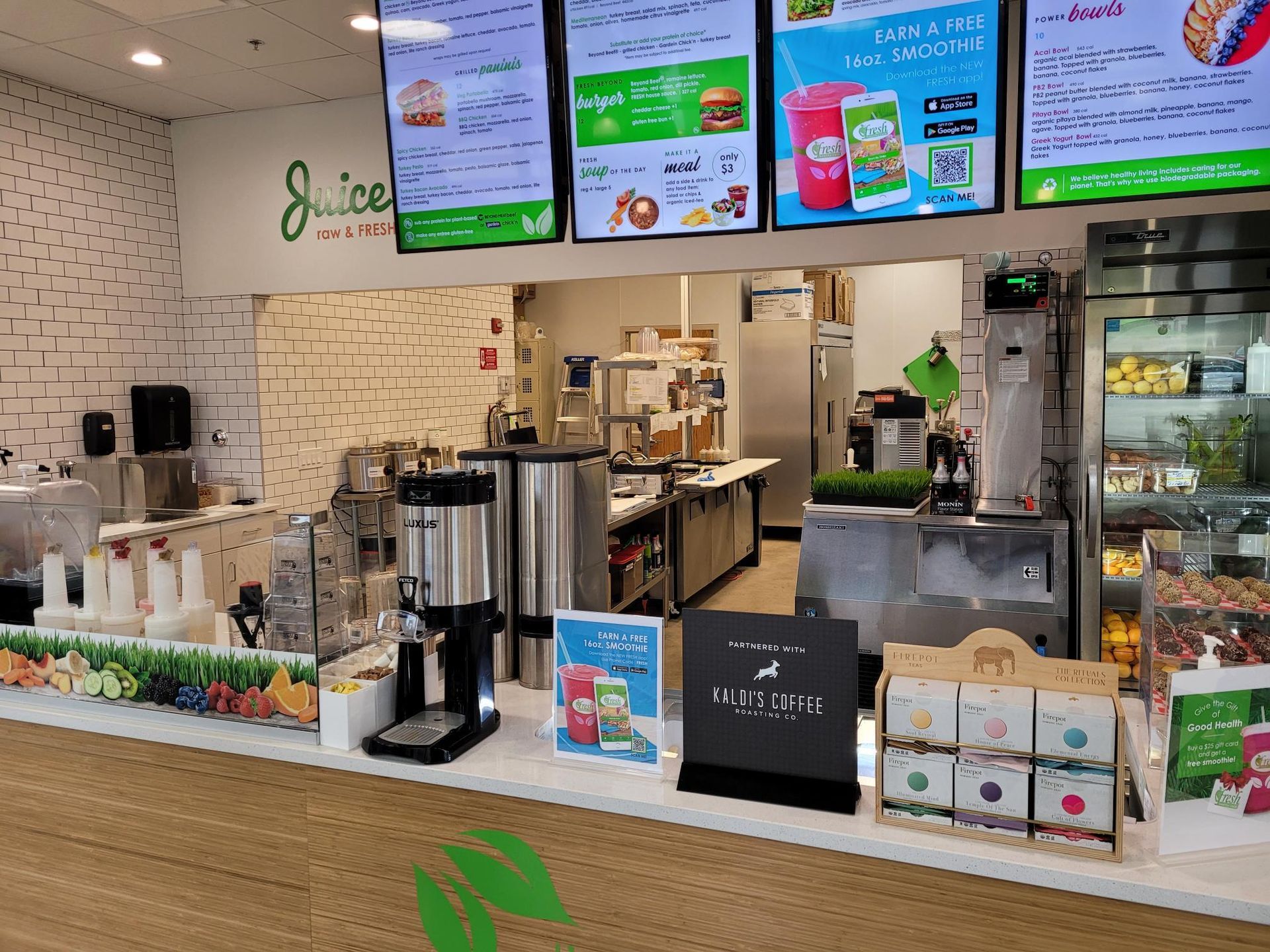 A brightly lit juice bar interior with digital menu boards above a service counter stocked with supplies and products.