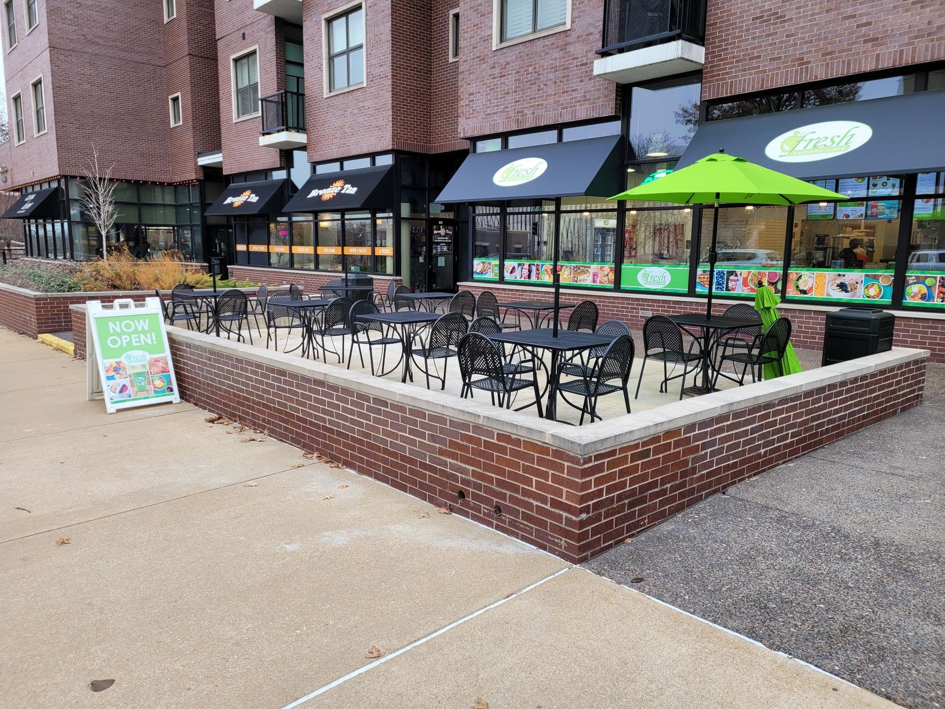 A street-level outdoor patio with black metal tables and chairs, a bright green umbrella, and a sidewalk sign for a cafe.