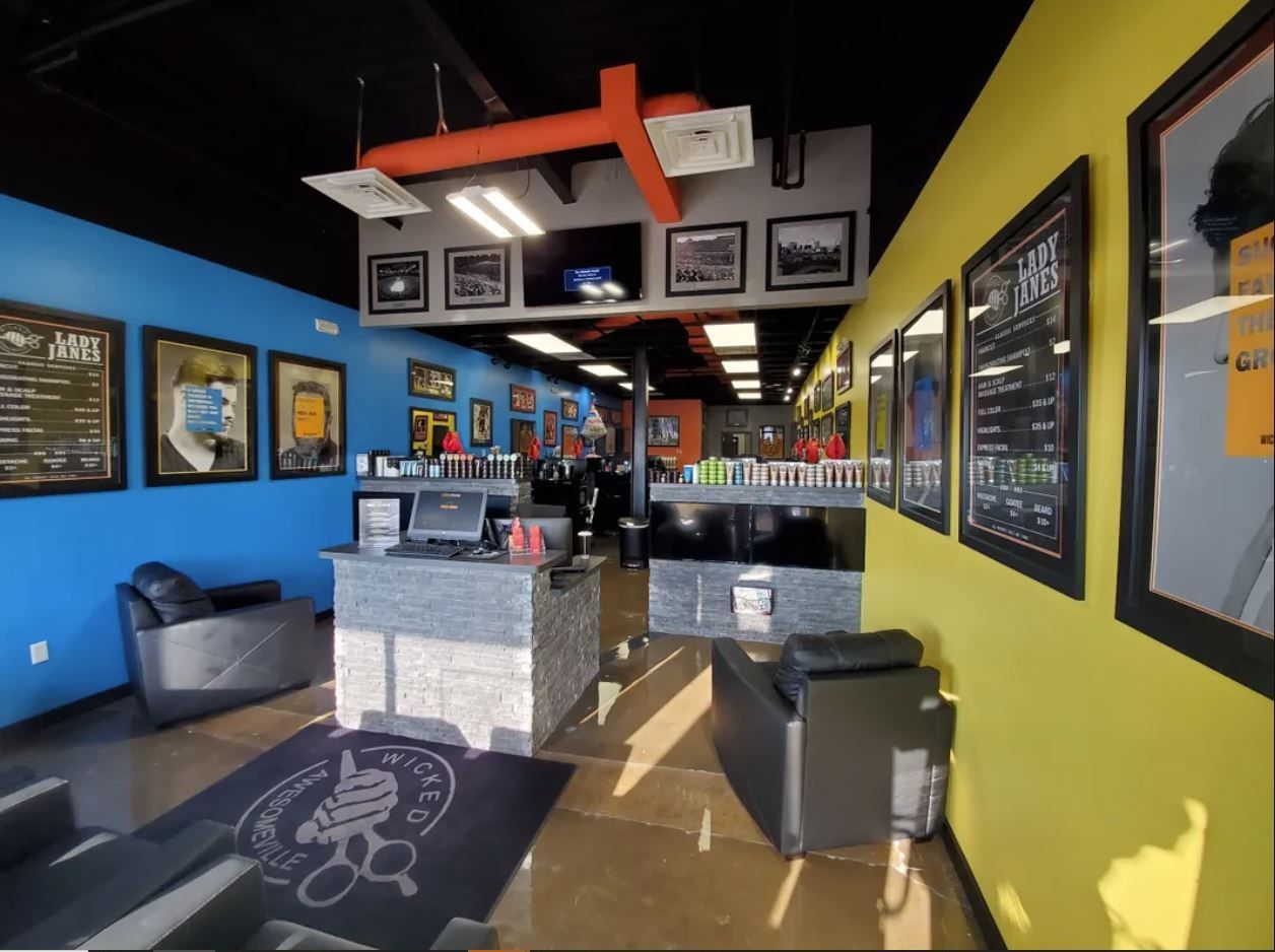 A brightly lit barbershop interior with a blue wall, yellow wall, reception desk, black lounge chairs, and wall art.