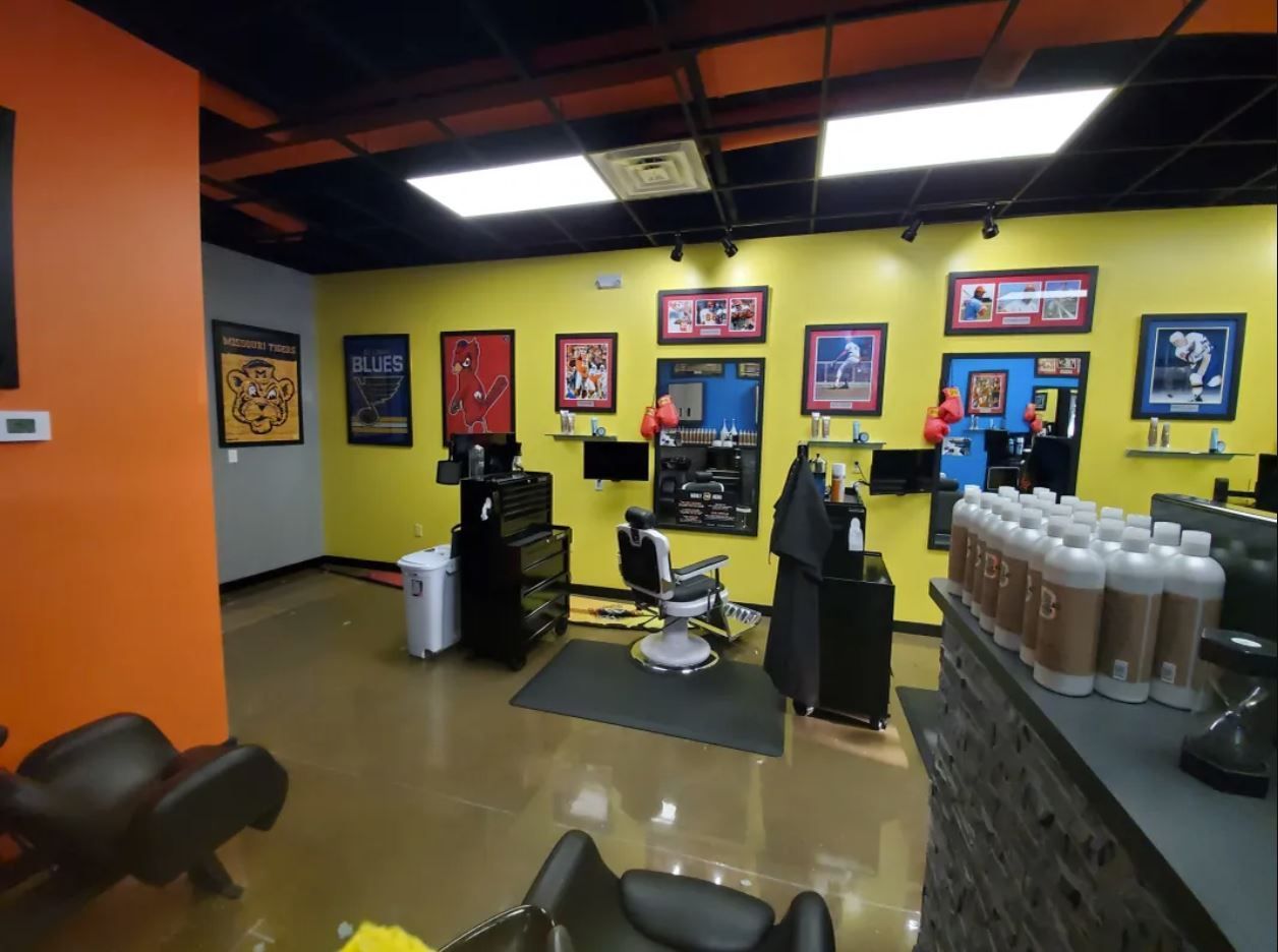 A modern, bright-yellow barber shop with salon chairs, framed sports posters on the walls, and product bottles on a counter.