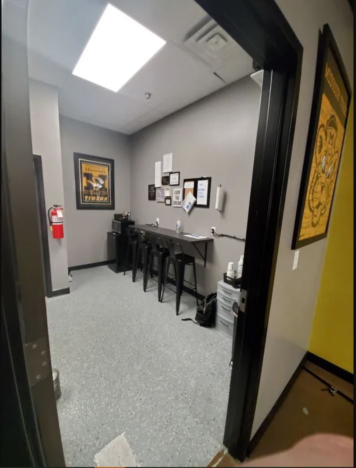 A small break room with gray walls, a high counter with three black barstools, wall art, and a fire extinguisher.