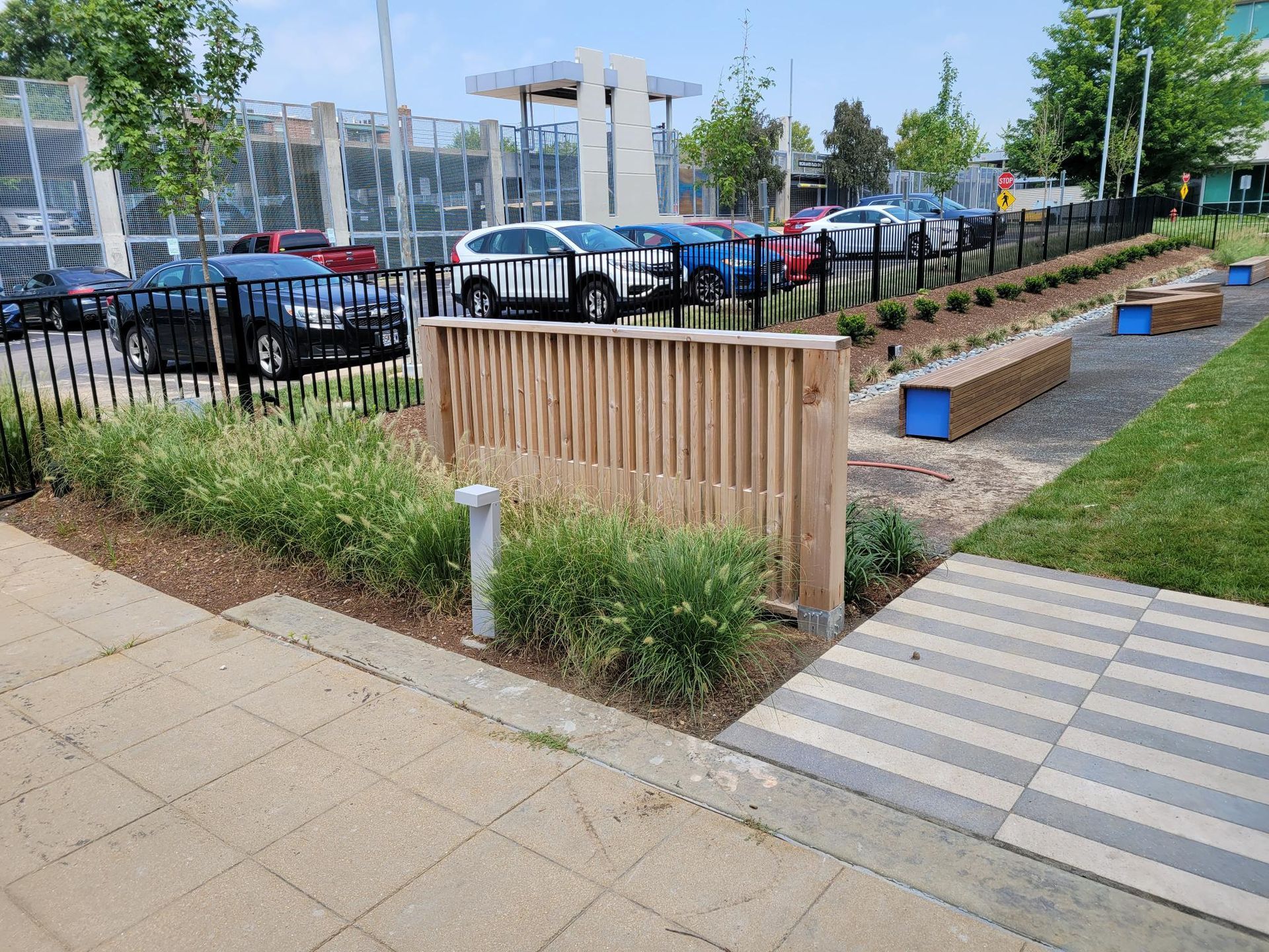 A paved walkway and lawn area with wooden benches, a slat partition, and landscaping near a parking lot with several cars.