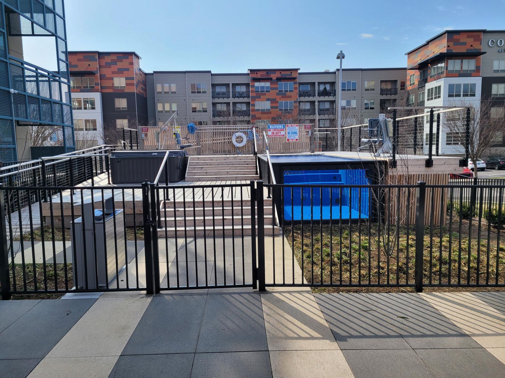A fenced-in outdoor area with a blue-tiled swimming pool and a hot tub, set against modern multi-story apartment buildings.