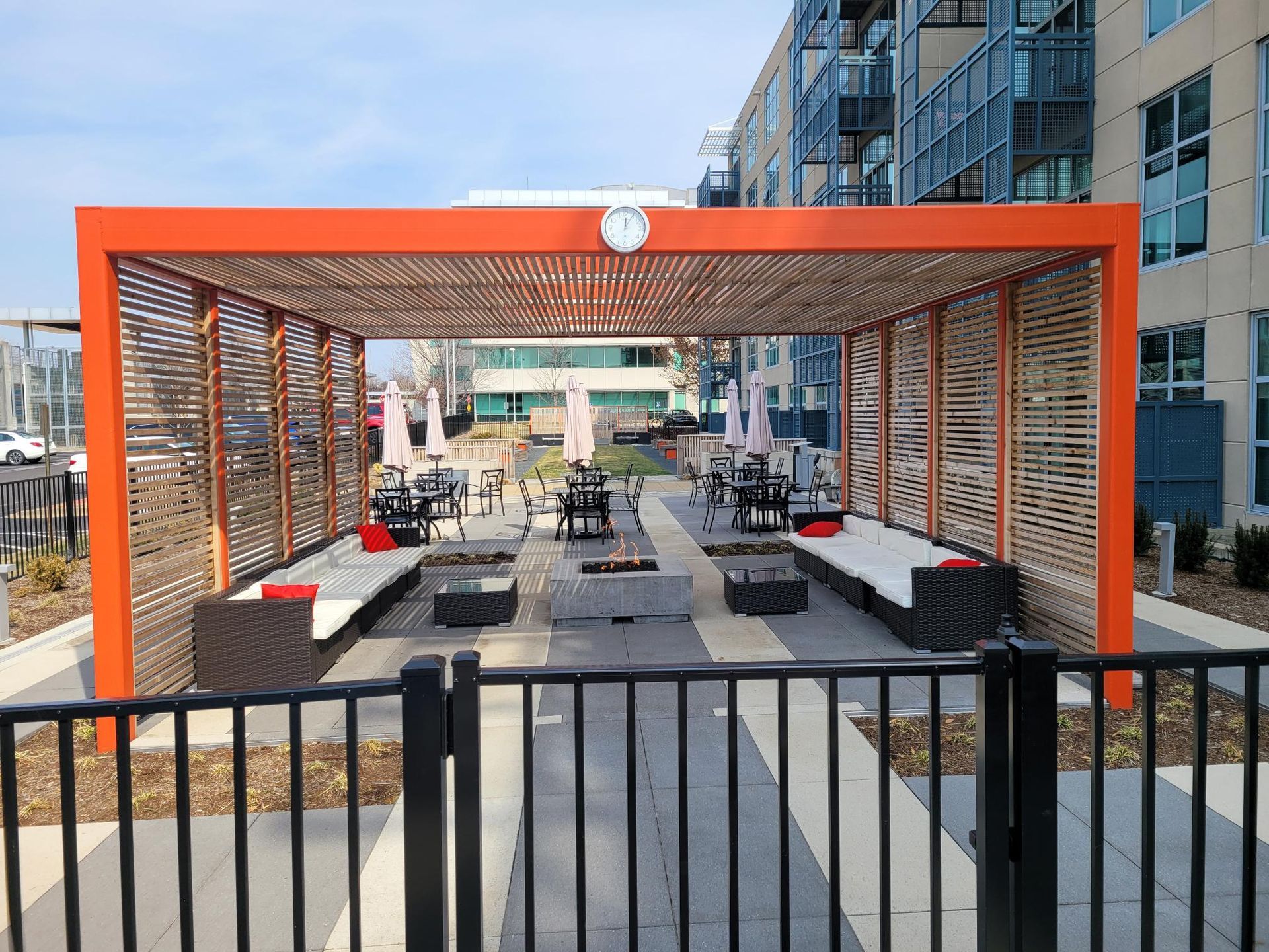 An orange-framed outdoor patio structure with wooden slats, featuring lounge furniture and fire pits on a tiled floor.
