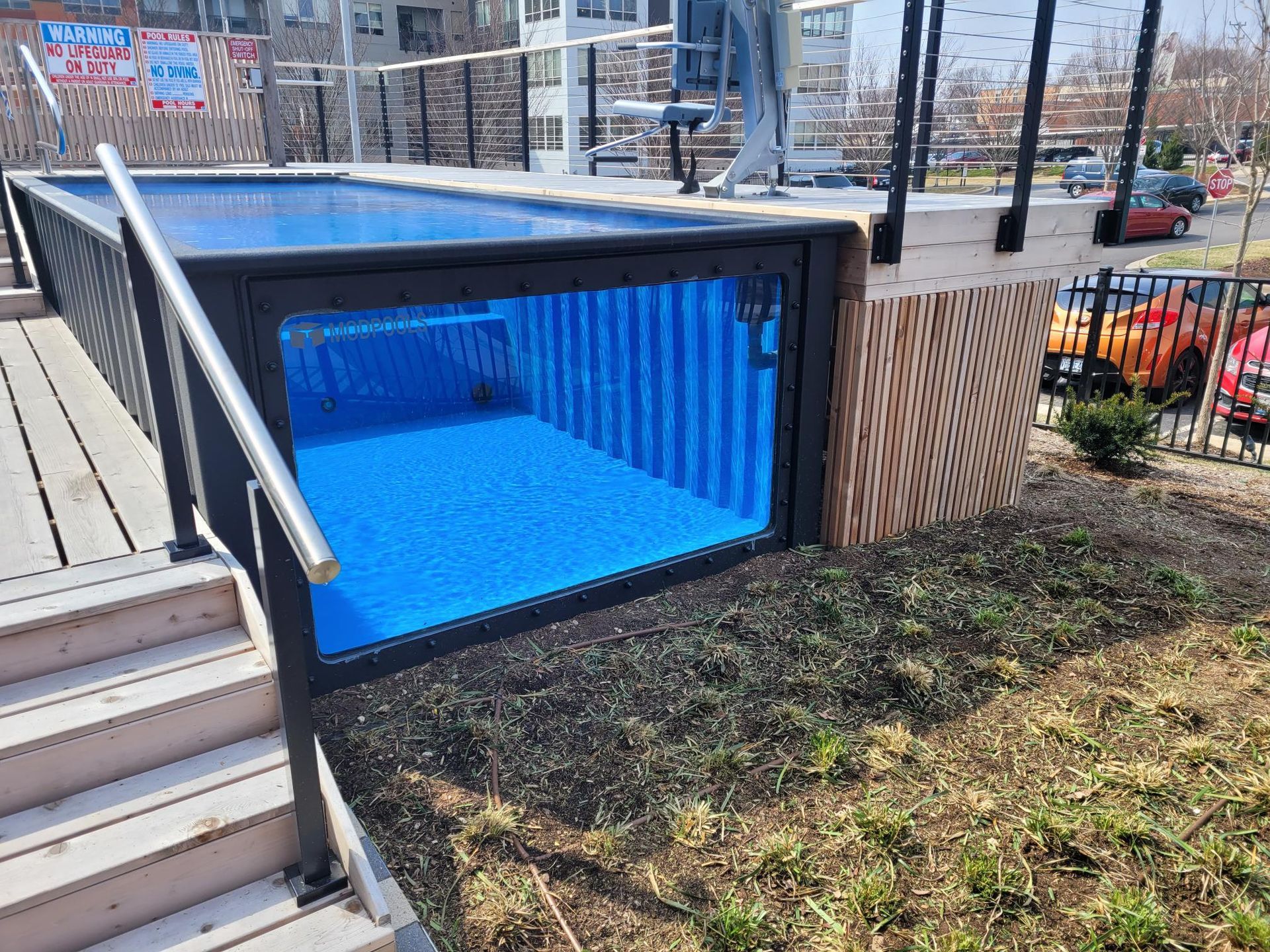 An above-ground pool made from a shipping container with a large glass side panel, situated next to stairs and a deck.