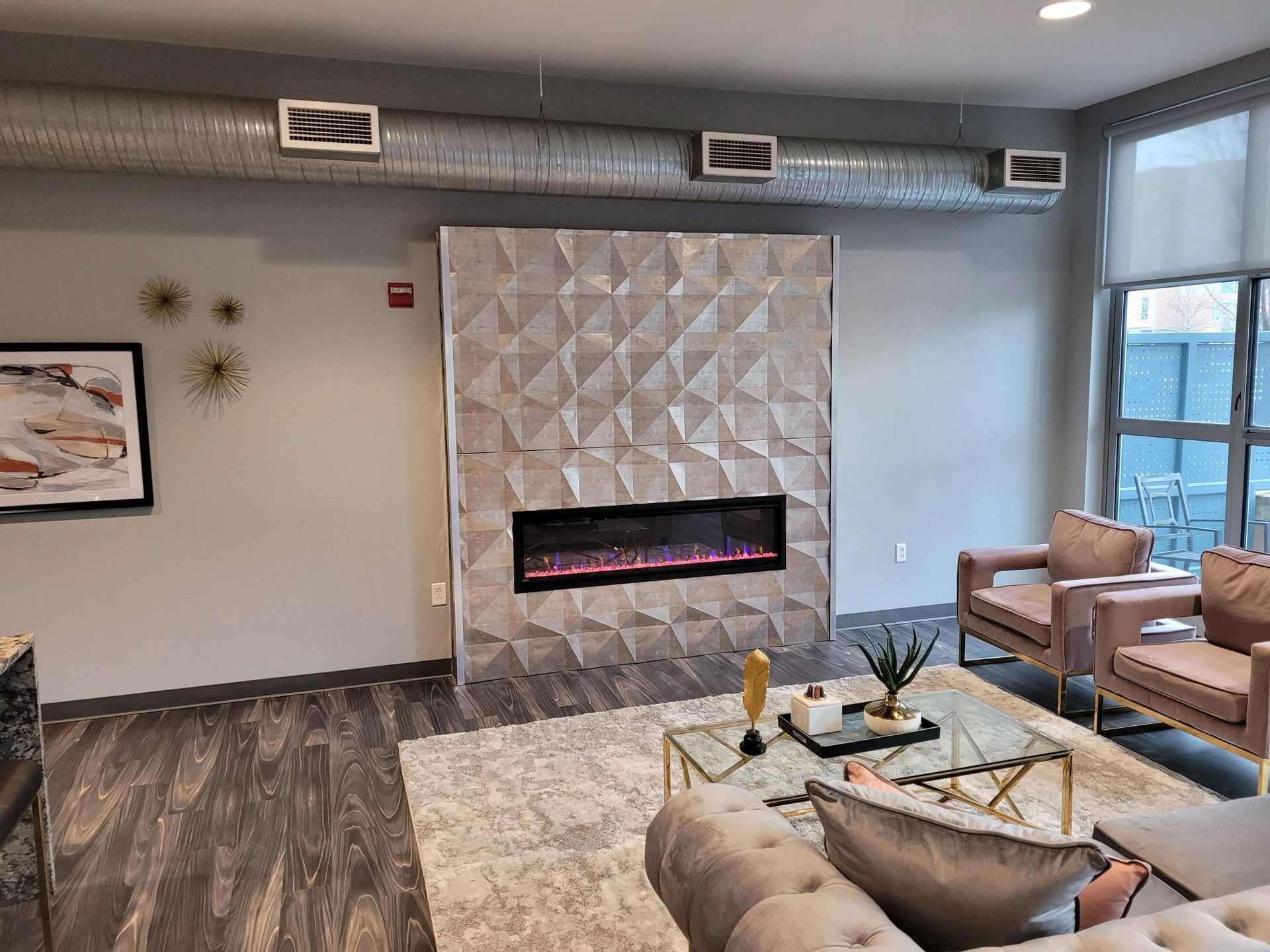 A modern living space features a fireplace with a geometric textured wall, beige seating, and exposed ductwork.