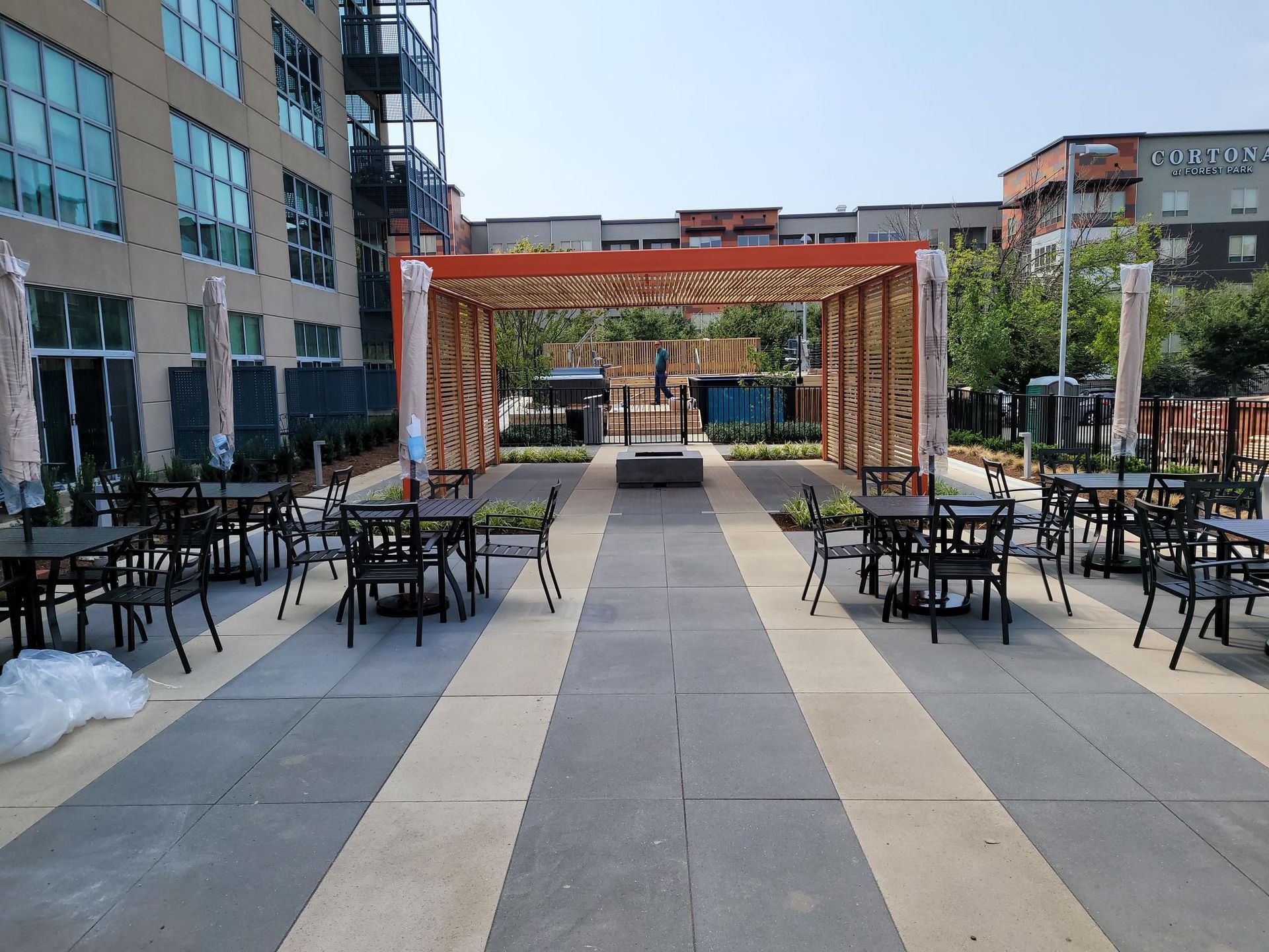 Outdoor patio with cafe tables and chairs under a decorative orange pergola on a paved walkway between multi-story buildings.