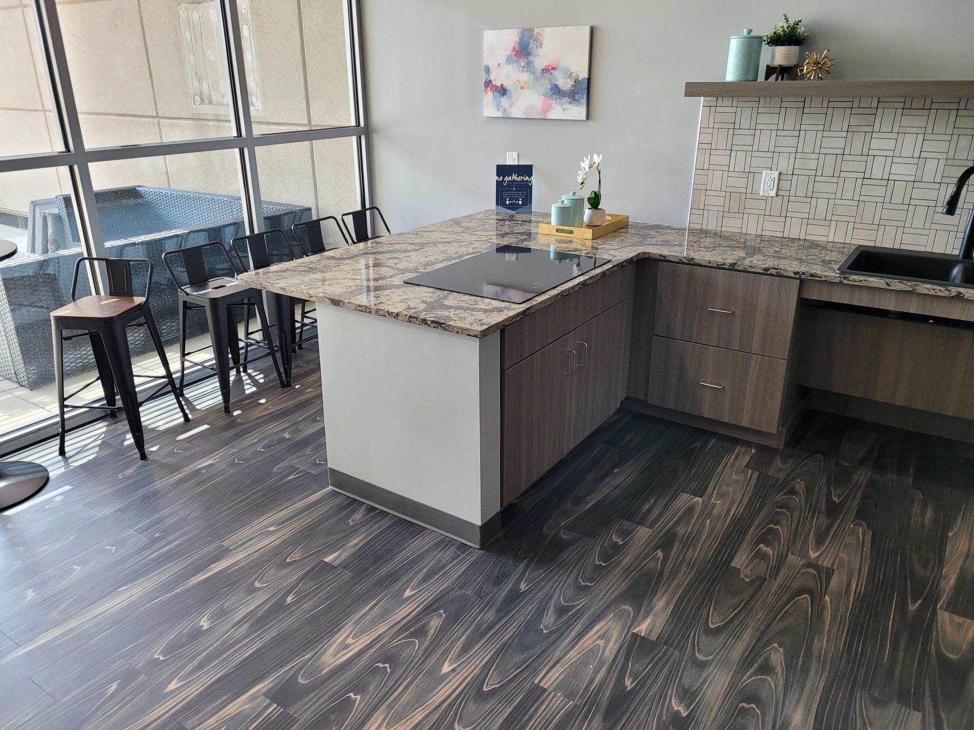 A modern kitchenette with a granite island, dark wood cabinets, and stools, positioned near a large window.