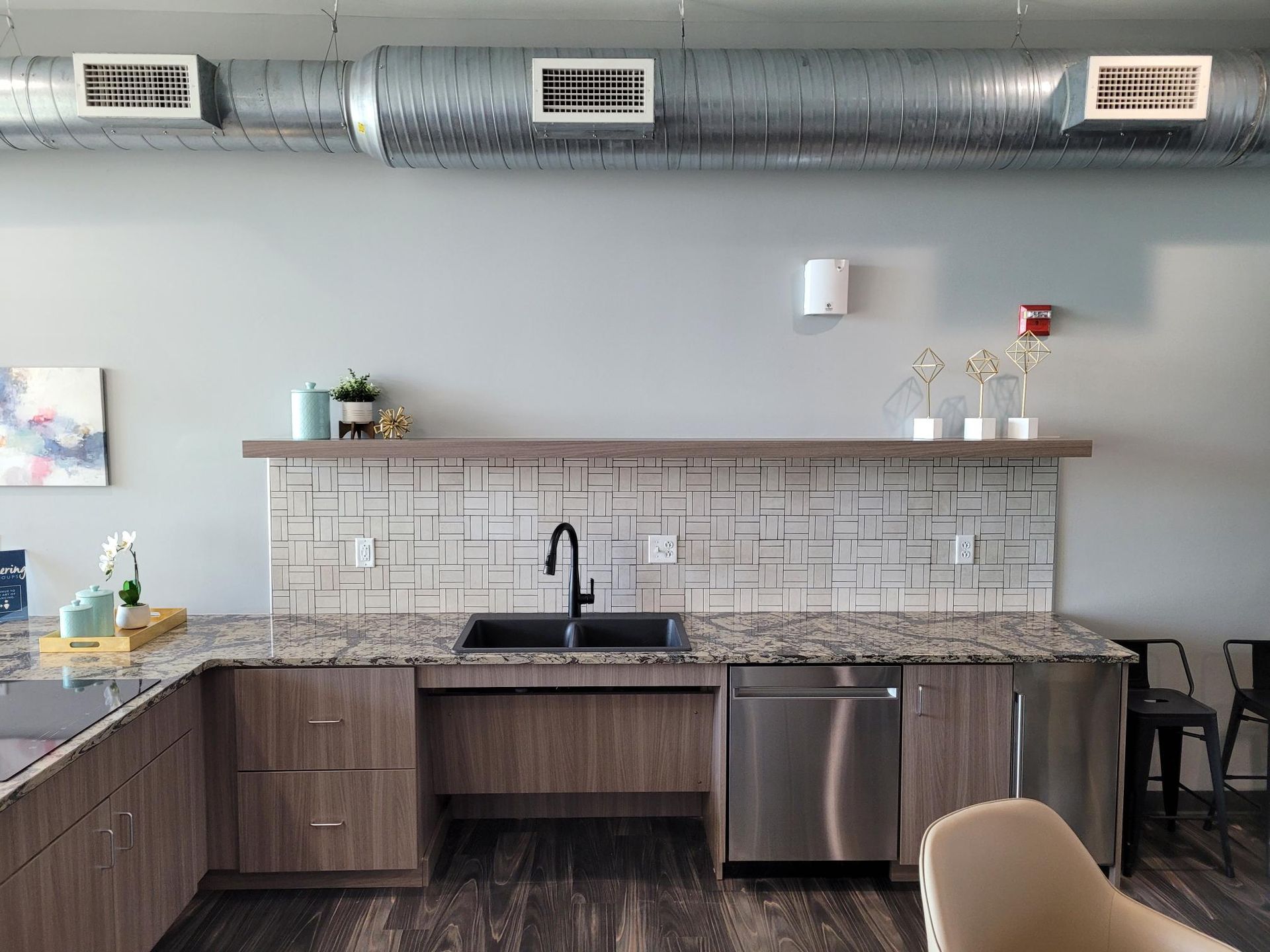 A modern kitchen area with brown cabinets, granite countertops, a tiled backsplash, and exposed ductwork above.