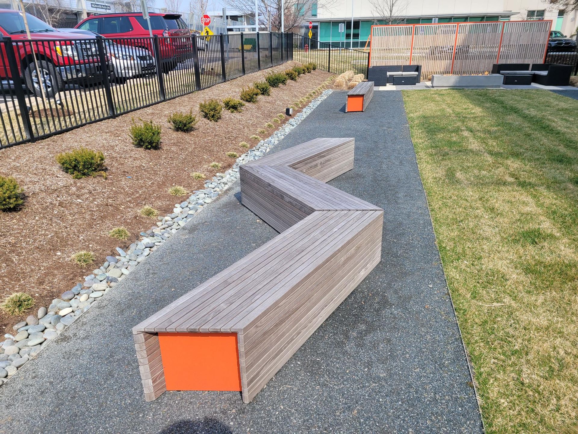 An outdoor zigzag-shaped wooden bench with orange ends sits on a gravel path next to a lawn and a black metal fence.