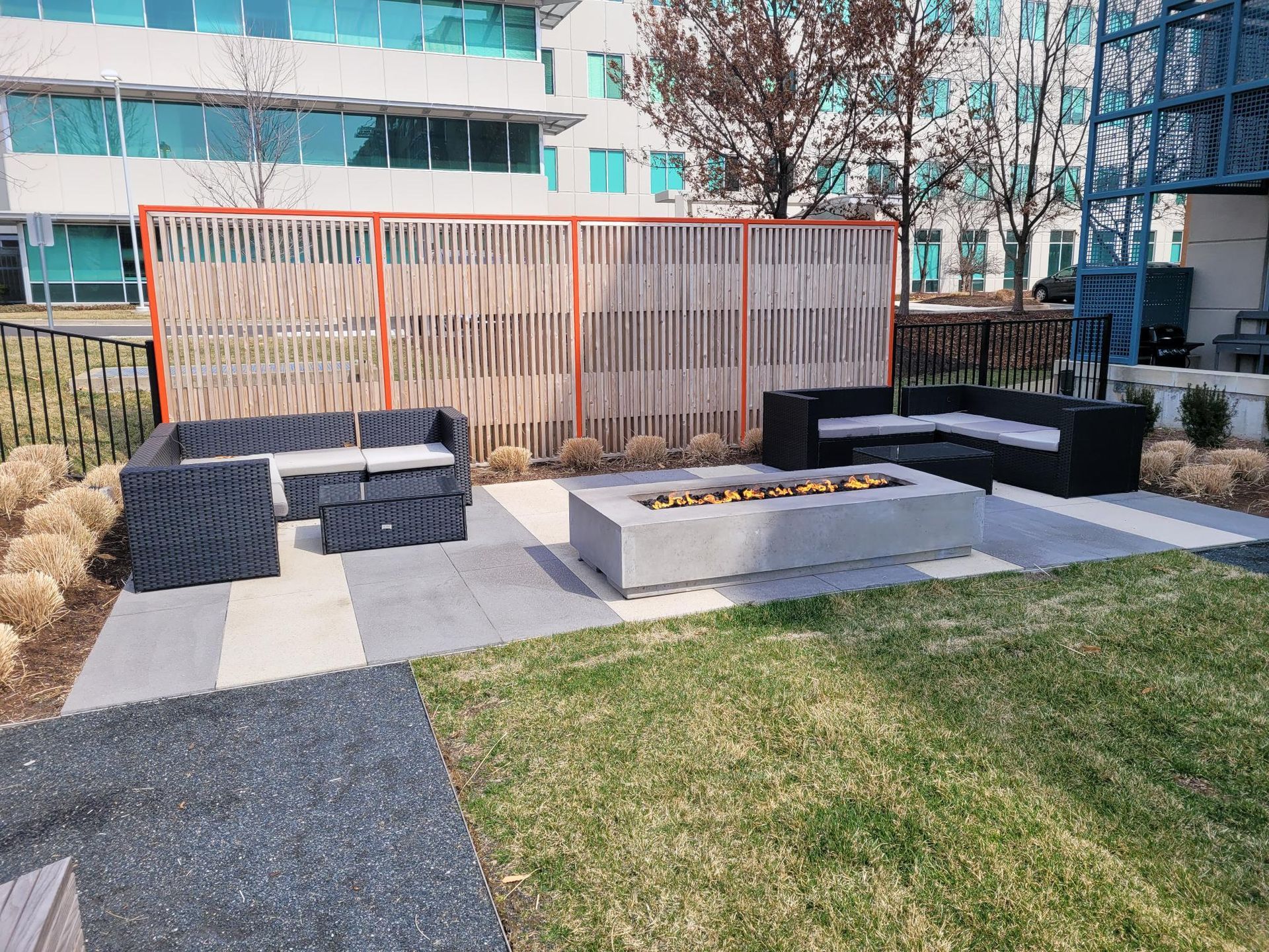Outdoor patio area featuring two dark wicker sofas and a long concrete fire pit on a tiled deck in front of a wood fence.