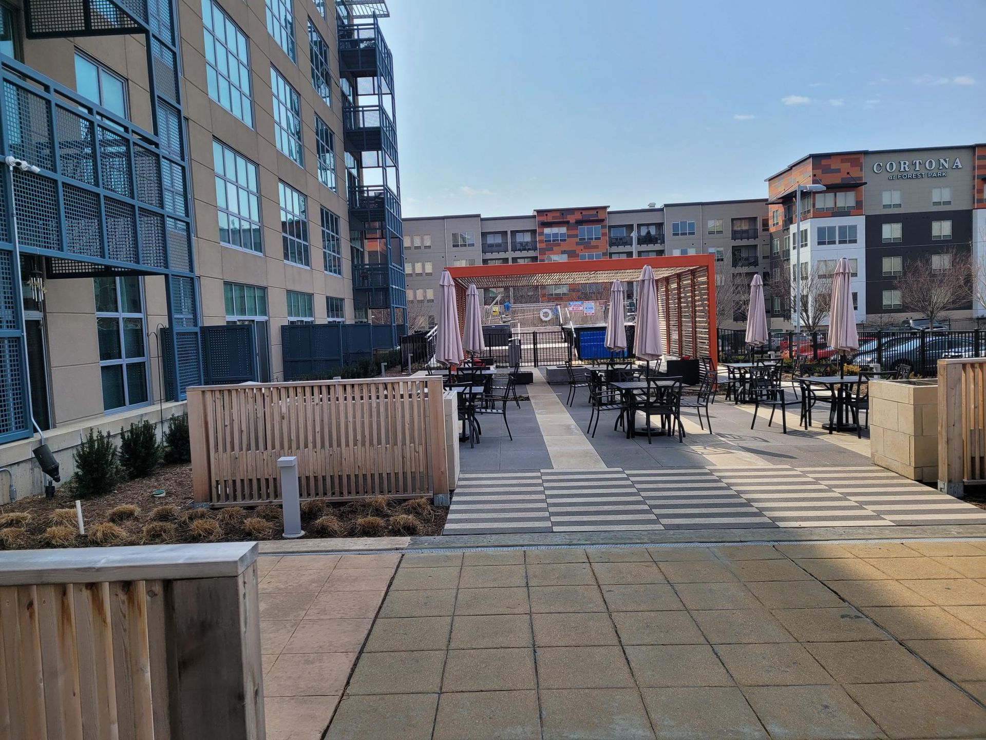 An outdoor patio space with tables, chairs, and a shaded pergola structure, bordered by modern apartment buildings.