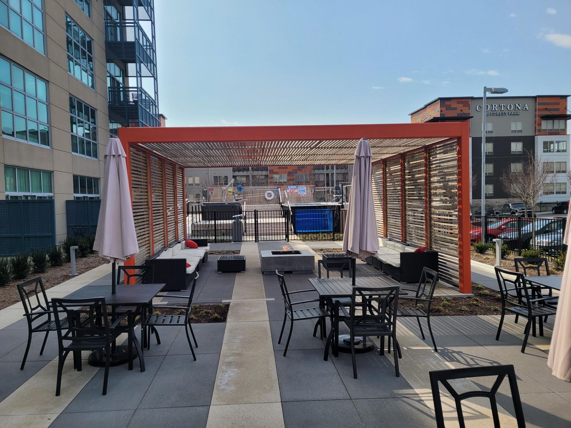 An outdoor patio with metal tables and chairs under a modern orange pergola, with a lounge area and stone fire pits.