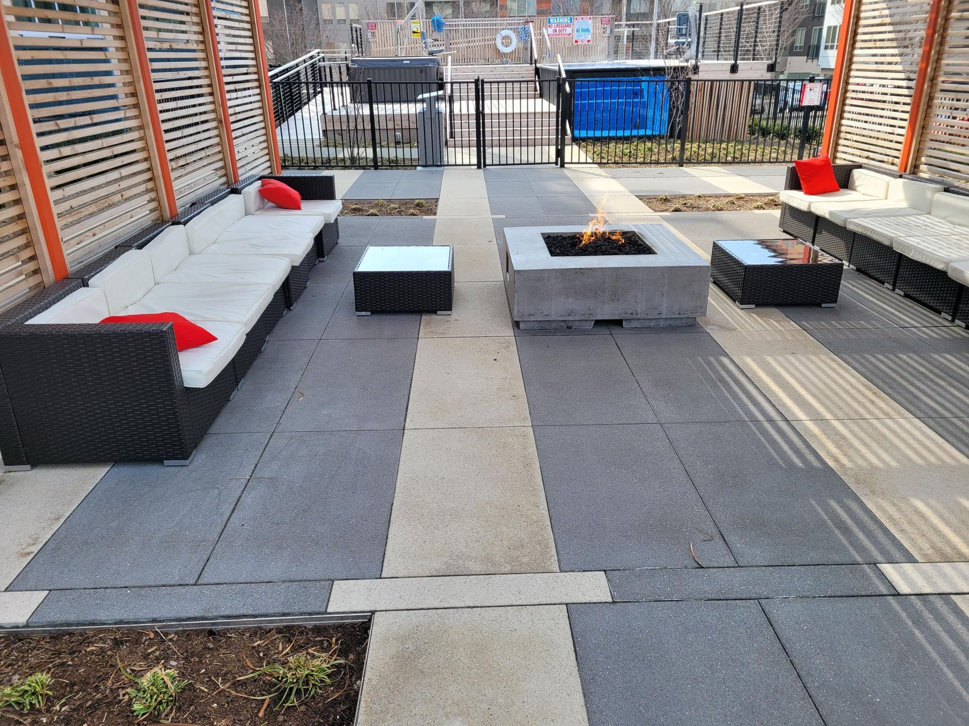 Outdoor patio with fire pit, modern black wicker seating, white cushions, and red throw pillows on a paved terrace.
