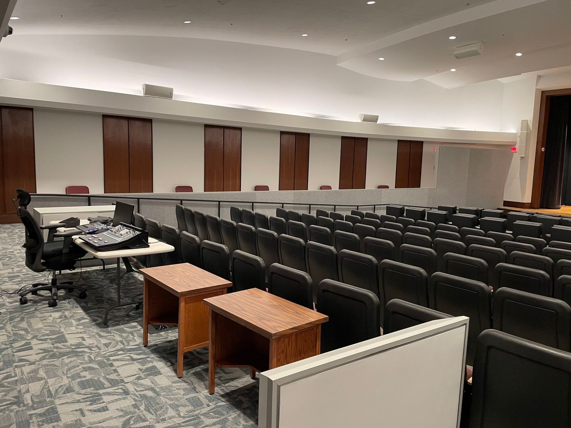 An empty auditorium with rows of black chairs facing a dais and a desk with audio equipment on the left.