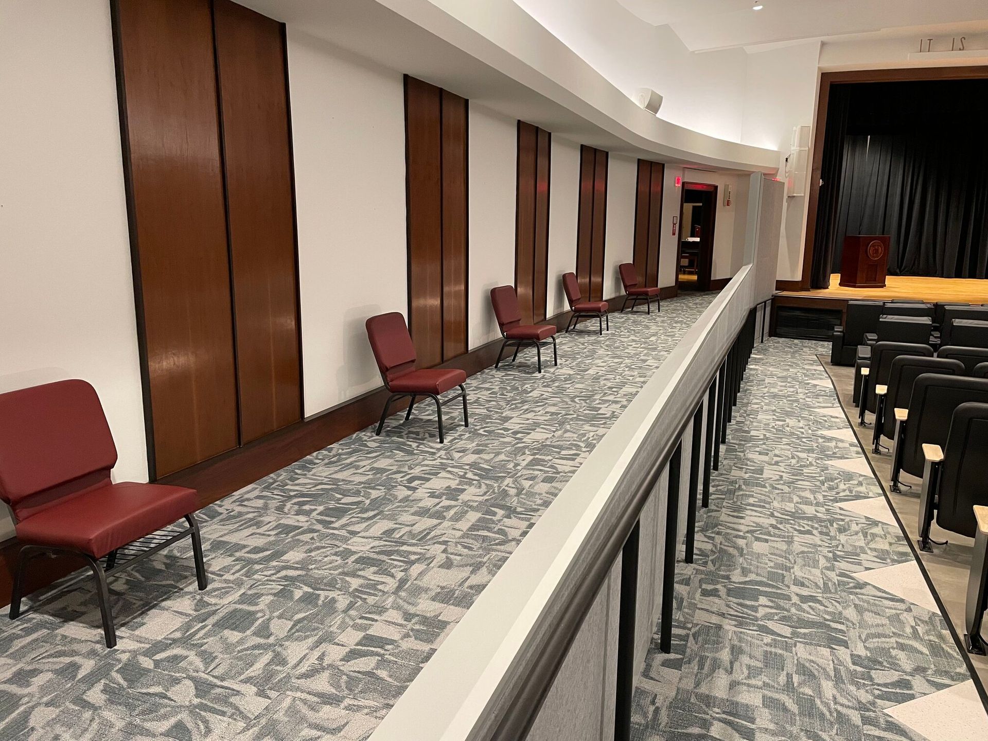 Rows of red chairs line a patterned carpet in a room with wood-paneled walls and a stage area.