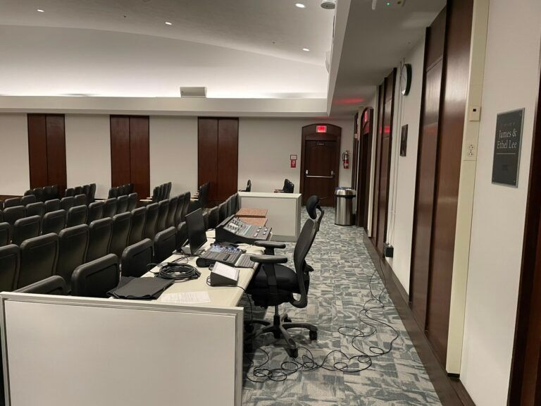 A lecture hall featuring rows of black chairs, a workstation with audio equipment, and wood-paneled walls.